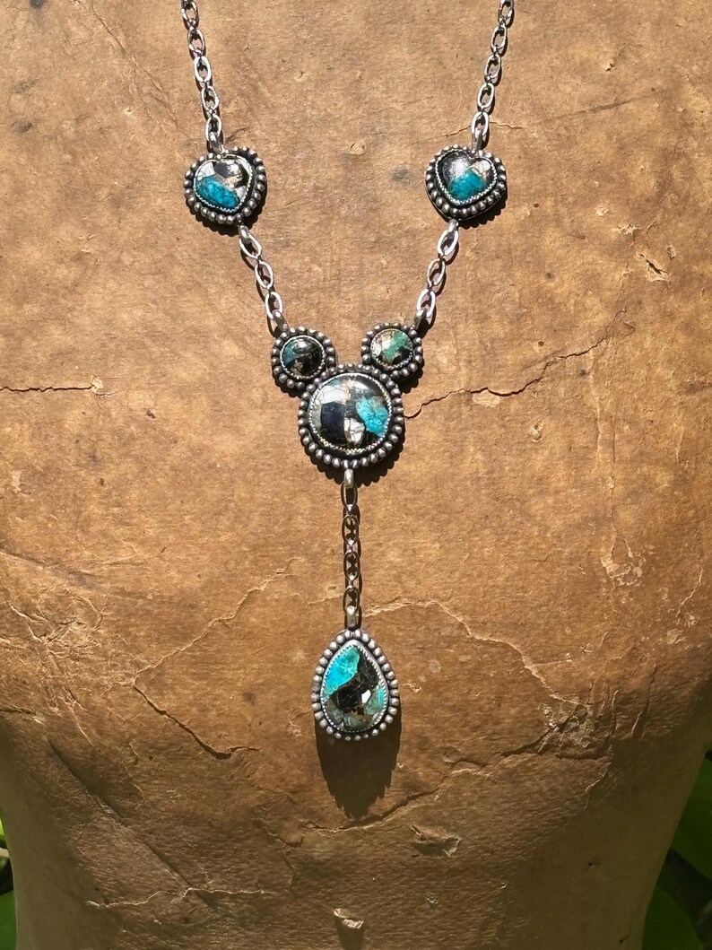 May include: A silver necklace featuring five bezel-set stones with turquoise and black matrix patterns. The design includes two heart-shaped stones, two small circular stones, one large central circular stone, and a dangling teardrop-shaped stone.