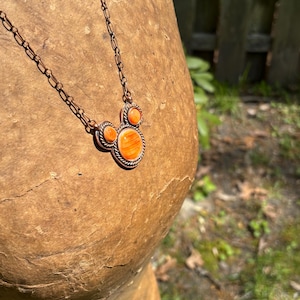 May include: A copper-toned necklace featuring a pendant with an orange, Mickey Mouse-shaped stone. The pendant is framed with a decorative border and is connected to a delicate chain. The necklace is displayed on a textured, brown surface.