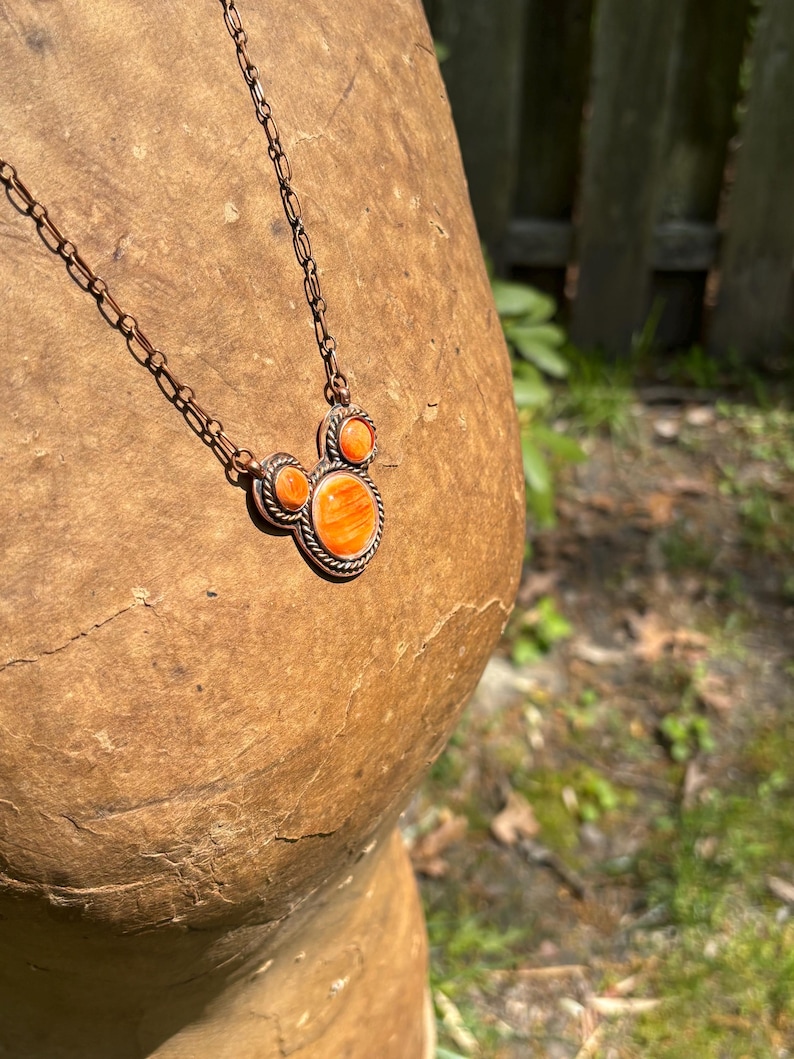 May include: A copper-toned necklace featuring a pendant with an orange, Mickey Mouse-shaped stone. The pendant is framed with a decorative border and is connected to a delicate chain. The necklace is displayed on a textured, brown surface.