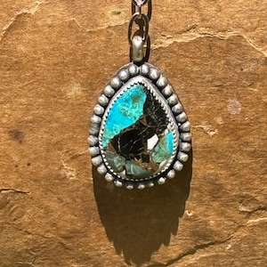 May include: A teardrop-shaped pendant featuring a multi-colored stone with vibrant turquoise and deep black matrix patterns. The stone is set in a silver bezel decorated with a beaded border and hangs from a silver chain link necklace.