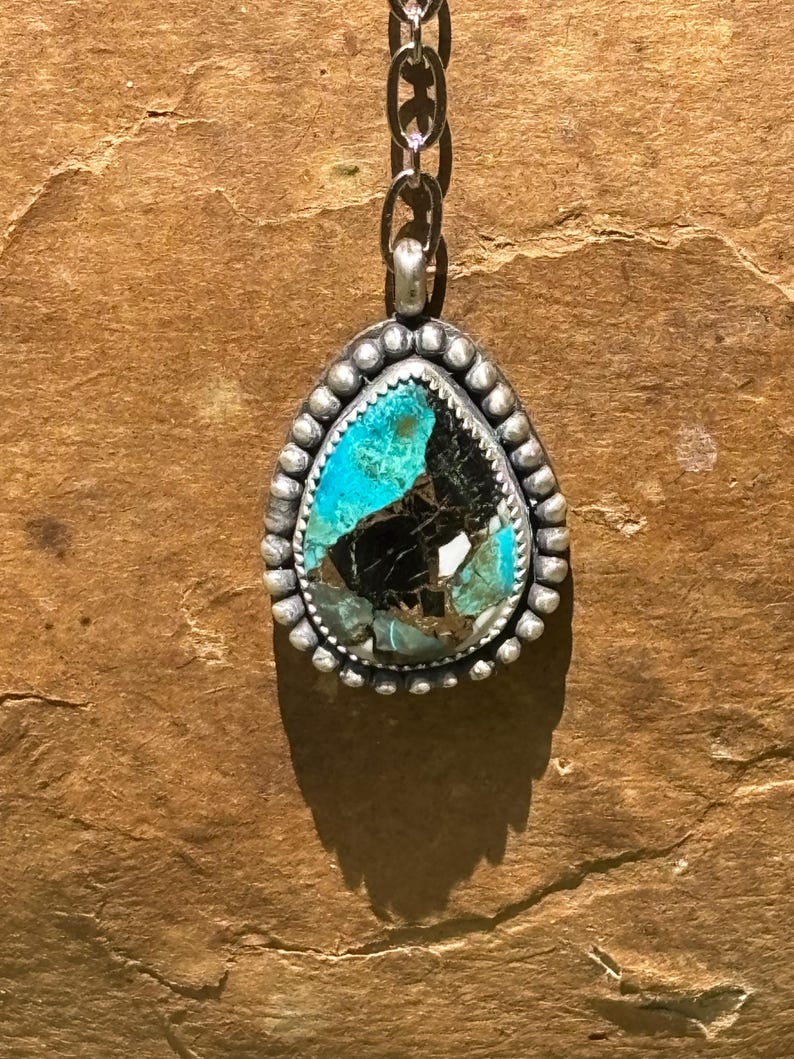 May include: A teardrop-shaped pendant featuring a multi-colored stone with vibrant turquoise and deep black matrix patterns. The stone is set in a silver bezel decorated with a beaded border and hangs from a silver chain link necklace.