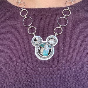 May include: A silver necklace with a pendant shaped like a mouse head. The pendant has a large, oval turquoise stone and two smaller stones. The necklace chain is made of silver rings.