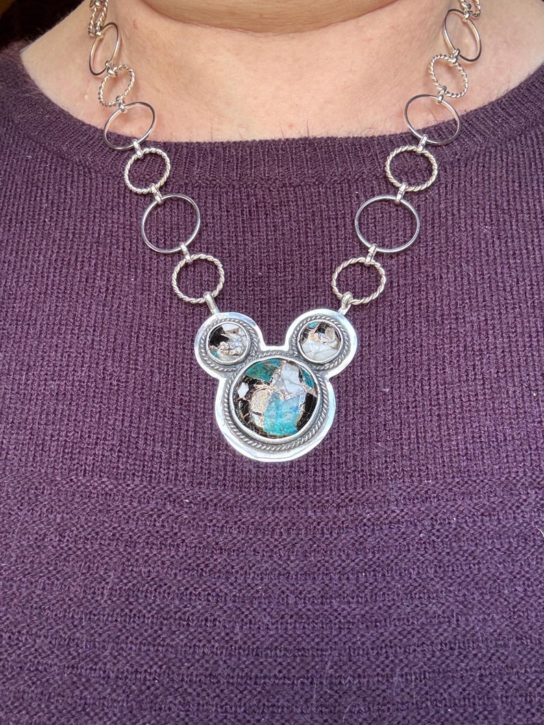 May include: A silver necklace with a pendant shaped like a mouse head. The pendant has a large, oval turquoise stone and two smaller stones. The necklace chain is made of silver rings.