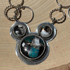 May include: Silver necklace with a pendant shaped like a mouse head, featuring three circular stone settings. The central setting has a turquoise, black, white, and gold stone. The necklace has a series of silver rings.