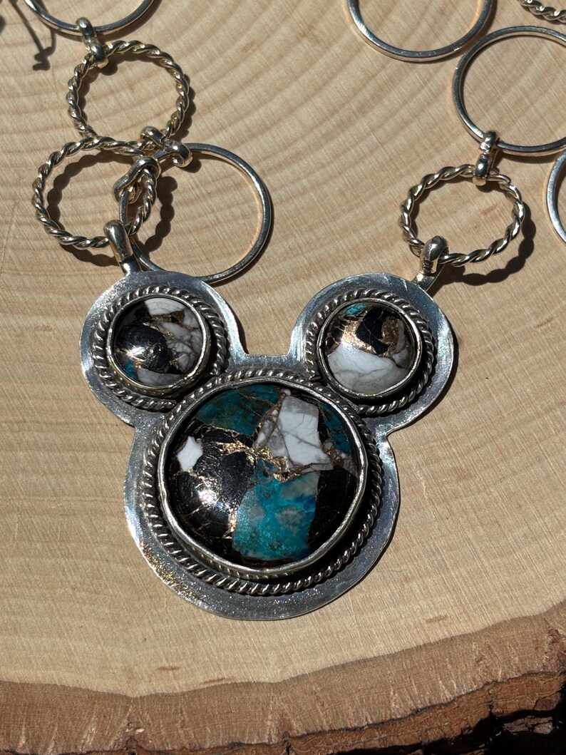 May include: Silver necklace with a pendant shaped like a mouse head, featuring three circular stone settings. The central setting has a turquoise, black, white, and gold stone. The necklace has a series of silver rings.