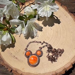 May include: A copper-toned necklace with an orange stone pendant in a Mickey Mouse-inspired design. The necklace is displayed on a wooden slice, with white flowers and green leaves in the background.