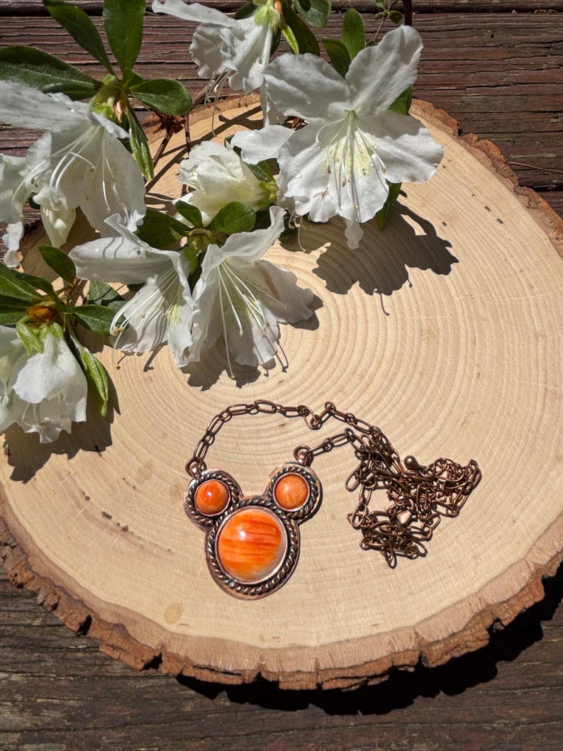 May include: A copper-toned necklace with an orange stone pendant in a Mickey Mouse-inspired design. The necklace is displayed on a wooden slice, with white flowers and green leaves in the background.