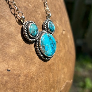Pu&ograve; includere: Una collana d'argento con tre pietre turchesi. Le pietre ovali sono incastonate in un bordo argentato a forma di corda. La catena &egrave; una delicata maglia d'argento. La collana &egrave; esposta su una superficie di legno marrone.