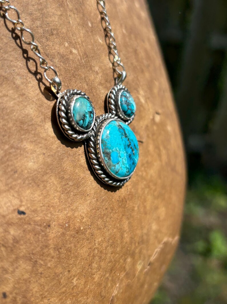 Pu&ograve; includere: Una collana d'argento con tre pietre turchesi. Le pietre ovali sono incastonate in un bordo argentato a forma di corda. La catena &egrave; una delicata maglia d'argento. La collana &egrave; esposta su una superficie di legno marrone.