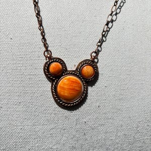 May include: A copper-colored necklace with three round, orange-colored stone pendants. The pendants are set in a decorative, rope-like border. The necklace chain is also copper-colored.