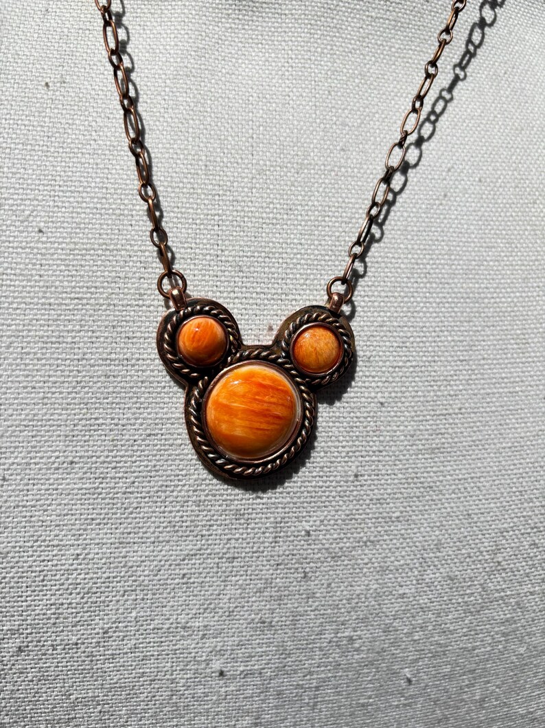 May include: A copper-colored necklace with three round, orange-colored stone pendants. The pendants are set in a decorative, rope-like border. The necklace chain is also copper-colored.
