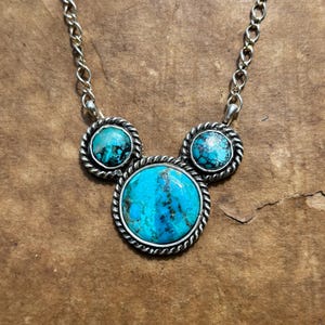 Pu&ograve; includere: Collana color argento con tre pietre turchesi. Le pietre sono incastonate in una cornice d'argento con un design a corda ritorta. La collana ha una catena d'argento.