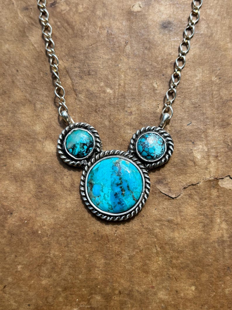 Pu&ograve; includere: Collana color argento con tre pietre turchesi. Le pietre sono incastonate in una cornice d'argento con un design a corda ritorta. La collana ha una catena d'argento.