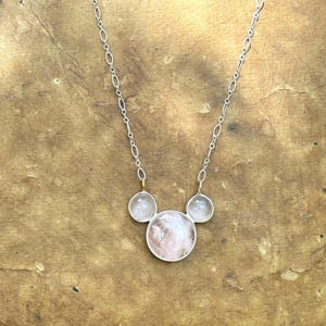 Puede incluir: Un collar de plata con tres piedras de cuarzo rosa. La piedra central es m&aacute;s grande y redonda, flanqueada por dos piedras redondas m&aacute;s peque&ntilde;as. La cadena del collar es delicada y plateada, con un dise&ntilde;o de eslabones ovalados. Las piedras tienen un tono rosa suave.