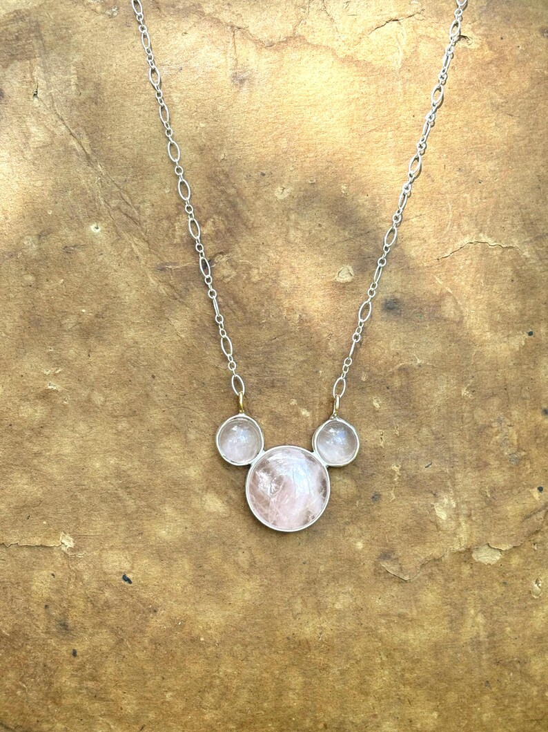 Puede incluir: Un collar de plata con tres piedras de cuarzo rosa. La piedra central es m&aacute;s grande y redonda, flanqueada por dos piedras redondas m&aacute;s peque&ntilde;as. La cadena del collar es delicada y plateada, con un dise&ntilde;o de eslabones ovalados. Las piedras tienen un tono rosa suave.