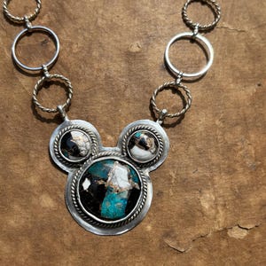 May include: A silver necklace with a pendant featuring three circular stone settings. The central setting has a large oval turquoise, black, and white stone. The smaller settings have similar stones. The necklace chain has silver rings and twisted rope accents.
