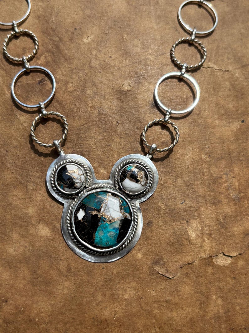 May include: A silver necklace with a pendant featuring three circular stone settings. The central setting has a large oval turquoise, black, and white stone. The smaller settings have similar stones. The necklace chain has silver rings and twisted rope accents.