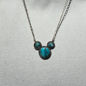 Pu&ograve; includere: Collana color argento con tre pietre turchesi. La pietra centrale, rotonda, &egrave; pi&ugrave; grande, affiancata da due pietre pi&ugrave; piccole. Le pietre turchesi sono incastonate in metallo argentato con un bordo a forma di corda. La catena &egrave; delicata.