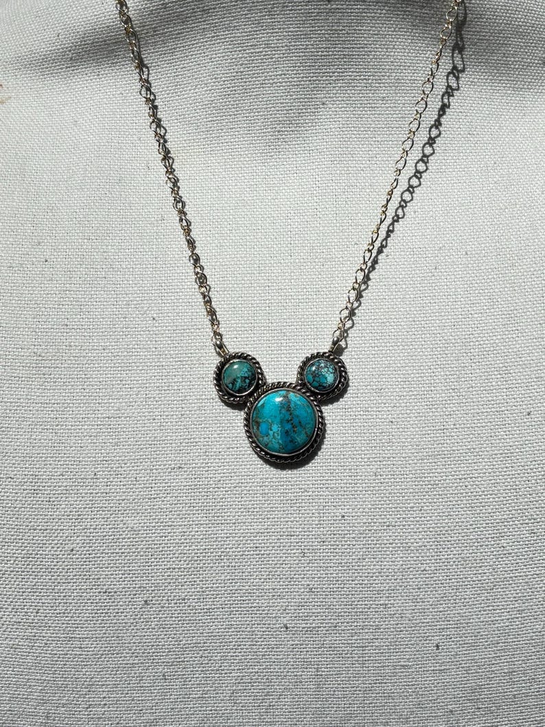 Pu&ograve; includere: Collana color argento con tre pietre turchesi. La pietra centrale, rotonda, &egrave; pi&ugrave; grande, affiancata da due pietre pi&ugrave; piccole. Le pietre turchesi sono incastonate in metallo argentato con un bordo a forma di corda. La catena &egrave; delicata.