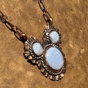 May include: A copper necklace featuring three oval blue lace agate stones set in scalloped and twisted wire bezels. The pendant is suspended from a thick copper chain with elongated oval links, showcasing a metallic finish and light blue banding.