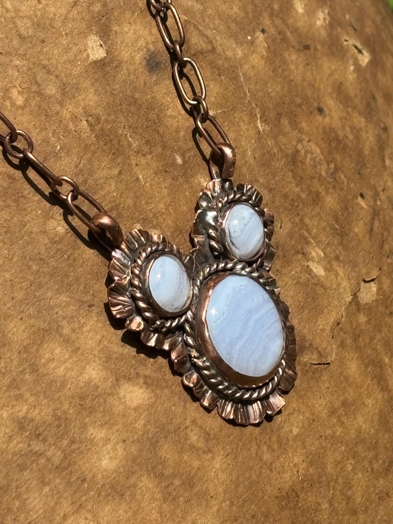May include: A copper necklace featuring three oval blue lace agate stones set in scalloped and twisted wire bezels. The pendant is suspended from a thick copper chain with elongated oval links, showcasing a metallic finish and light blue banding.