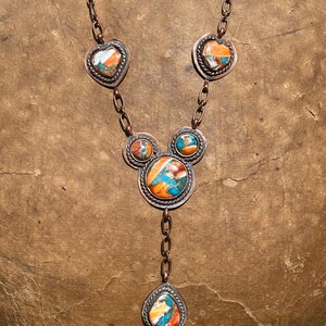 May include: A copper chain necklace featuring five bezel-set stones with marbled orange, blue, and white patterns. The stones are arranged in heart, circular, and teardrop shapes, each framed by a twisted metal border and connected by copper links.