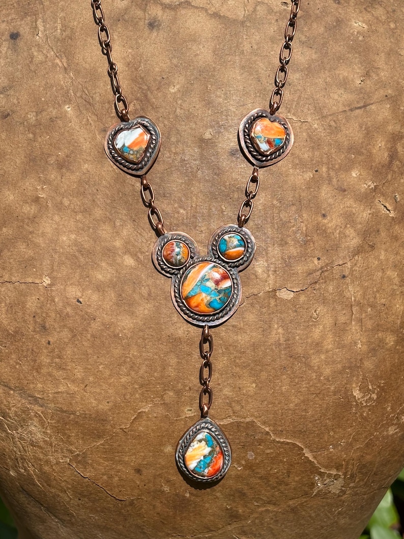 May include: A copper chain necklace featuring five bezel-set stones with marbled orange, blue, and white patterns. The stones are arranged in heart, circular, and teardrop shapes, each framed by a twisted metal border and connected by copper links.