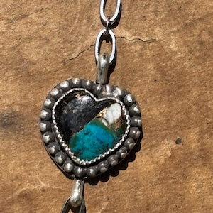 May include: A heart-shaped pendant featuring a dark stone with turquoise and white inclusions, set in a silver frame with a beaded edge. The pendant is attached to a silver chain with oval links, showcasing a textured, oxidized metal finish.