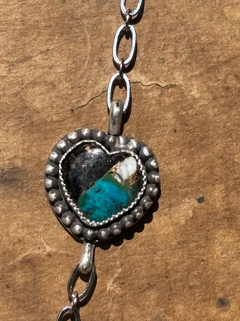 May include: A heart-shaped pendant featuring a dark stone with turquoise and white inclusions, set in a silver frame with a beaded edge. The pendant is attached to a silver chain with oval links, showcasing a textured, oxidized metal finish.