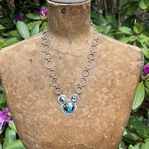 May include: A silver necklace with a pendant in a Mickey Mouse design. The pendant has a central circular stone with blue and black colours, surrounded by smaller circular accents. The necklace is displayed on a vintage mannequin.