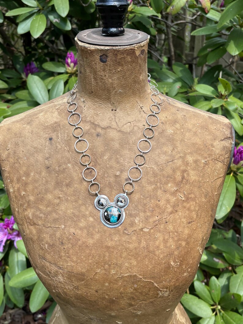 May include: A silver necklace with a pendant in a Mickey Mouse design. The pendant has a central circular stone with blue and black colours, surrounded by smaller circular accents. The necklace is displayed on a vintage mannequin.