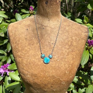 Pu&ograve; includere: Collana color argento con tre pietre turchesi. La pietra centrale &egrave; pi&ugrave; grande e rotonda, mentre le pietre laterali sono pi&ugrave; piccole. La collana &egrave; esposta su un manichino vintage, con uno sfondo di fogliame verde.
