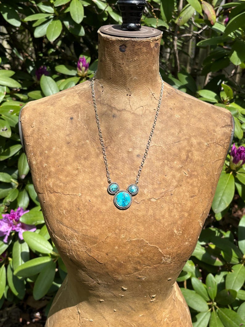 Pu&ograve; includere: Collana color argento con tre pietre turchesi. La pietra centrale &egrave; pi&ugrave; grande e rotonda, mentre le pietre laterali sono pi&ugrave; piccole. La collana &egrave; esposta su un manichino vintage, con uno sfondo di fogliame verde.