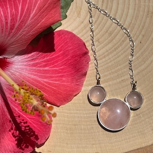 Puede incluir: Un collar de plata con tres piedras de cuarzo rosa, dispuestas en forma de cabeza de Mickey Mouse. El collar se exhibe sobre una superficie de madera, junto a una vibrante flor de hibisco rosa.