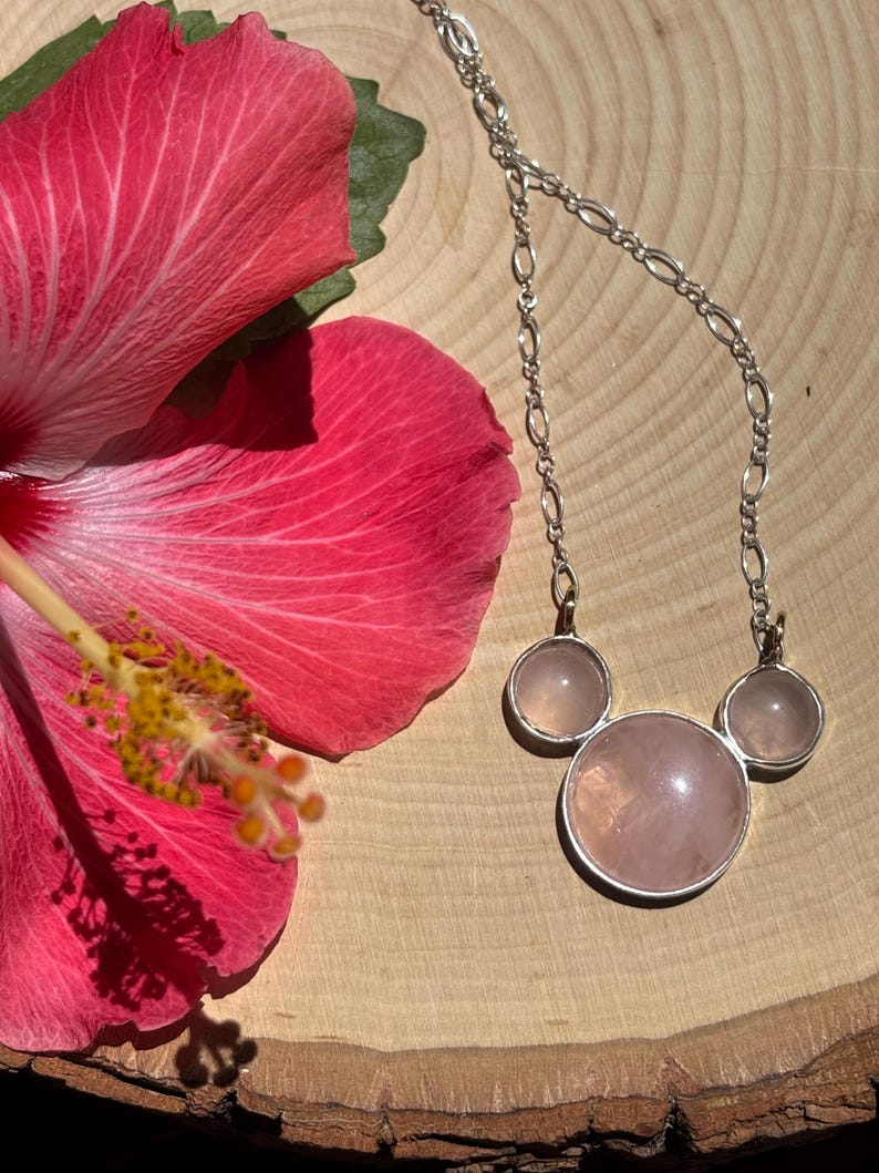 Puede incluir: Un collar de plata con tres piedras de cuarzo rosa, dispuestas en forma de cabeza de Mickey Mouse. El collar se exhibe sobre una superficie de madera, junto a una vibrante flor de hibisco rosa.