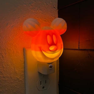 May include: A white and orange nightlight shaped like Mickey Mouse's head with a smiling face. The nightlight is plugged into an electrical outlet.