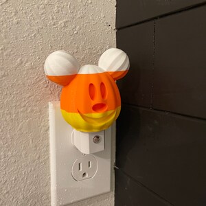 May include: A white night light shaped like Mickey Mouse with a candy corn design. The night light is plugged into a standard electrical outlet.