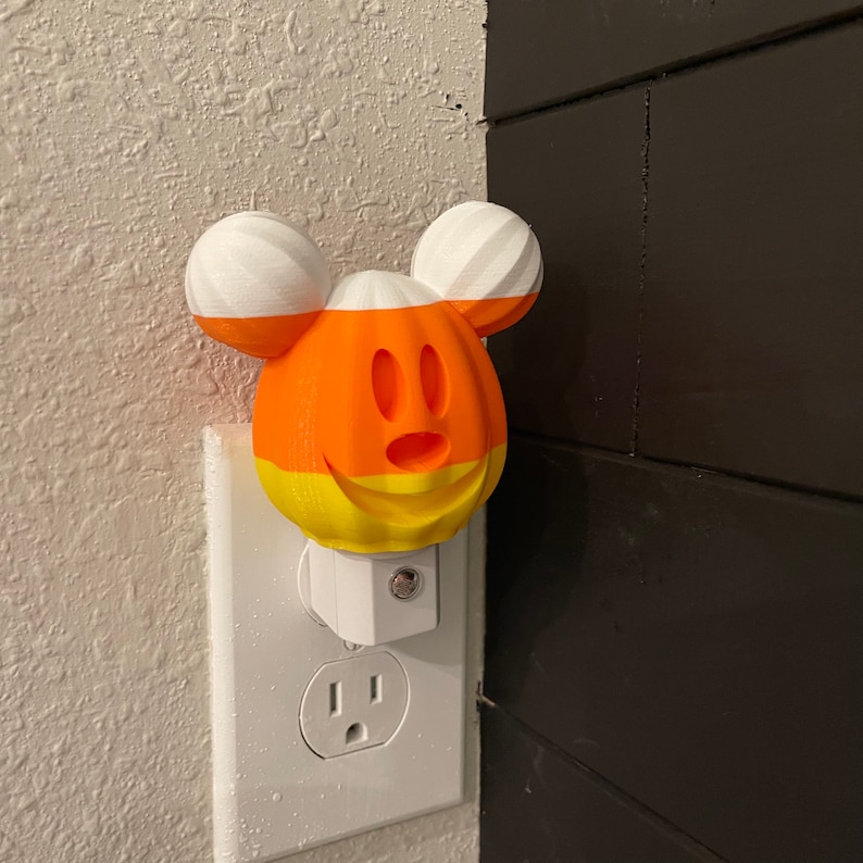 May include: A white night light shaped like Mickey Mouse with a candy corn design. The night light is plugged into a standard electrical outlet.