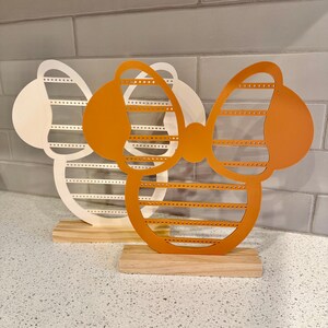 K&ouml;nnte beinhalten: Zwei Ohrringst&auml;nder in Form einer Schleife mit einem Holzsockel. Einer ist wei&szlig; und der andere orange.