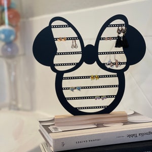 K&ouml;nnte beinhalten: Schwarzer Ohrringst&auml;nder aus Metall in Form eines Minnie Mouse-Kopfes mit Schleife. Der St&auml;nder hat mehrere Reihen von L&ouml;chern f&uuml;r Ohrringe. Der St&auml;nder steht auf einem Stapel B&uuml;cher mit dem Titel "Magnolia Table" von Joanna Gaines.