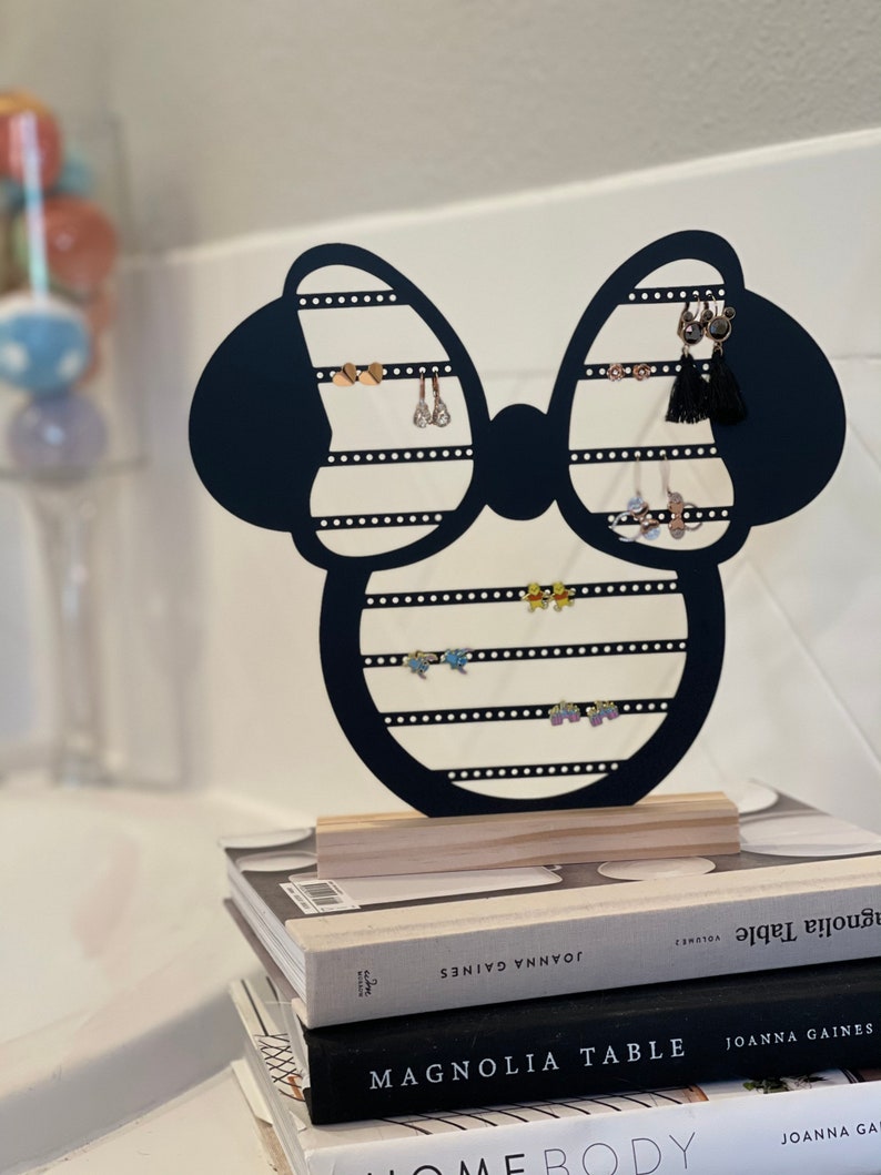 K&ouml;nnte beinhalten: Schwarzer Ohrringst&auml;nder aus Metall in Form eines Minnie Mouse-Kopfes mit Schleife. Der St&auml;nder hat mehrere Reihen von L&ouml;chern f&uuml;r Ohrringe. Der St&auml;nder steht auf einem Stapel B&uuml;cher mit dem Titel "Magnolia Table" von Joanna Gaines.