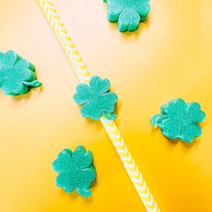 May include: Five green four-leaf clover shapes on a yellow background with a yellow and white striped straw.