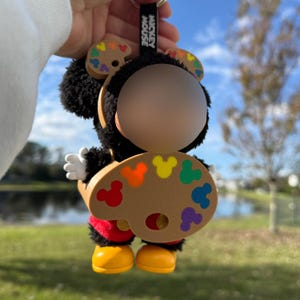 Könnte beinhalten: Ein Mickey Mouse Schlüsselanhänger mit einer Plüschfigur, die eine Malerpalette mit bunten Farbflecken hält. Die Figur hat schwarzes Fell, weiße Handschuhe, rote Shorts, gelbe Schuhe und die Aufschrift "Mickey Mouse" auf dem Schlüsselanhänger.