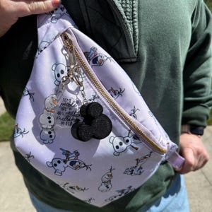 May include: A white fanny pack with a zipper closure and a gold zipper pull. The fanny pack has a repeating pattern of Olaf from the movie Frozen. The fanny pack has a black Mickey Mouse charm attached to it.