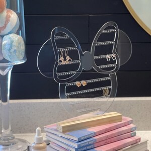 K&ouml;nnte beinhalten: Klarer Acryl-Ohrringst&auml;nder in Form einer Schleife mit einer Mickey-Maus-Kopf-Silhouette. Der St&auml;nder hat mehrere Reihen von L&ouml;chern f&uuml;r Ohrringe und steht auf einem Holzsockel.