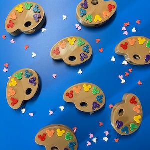 Könnte beinhalten: Ein Set aus acht bunten Farbpaletten in Form von Mickey-Maus-Köpfen. Jede Palette hat eine andere Farbe und ein Mickey-Maus-Kopf-Design.