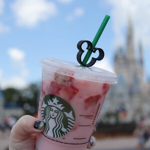 May include: A pink Starbucks drink with strawberry pieces in it. The drink has a Mickey Mouse straw topper and is being held in front of a blurred background of Cinderella's Castle at Walt Disney World.