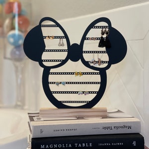 K&ouml;nnte beinhalten: Schwarzer Ohrringst&auml;nder aus Holz in Form einer Schleife mit Mickey-Maus-Ohren. Der St&auml;nder hat mehrere Reihen von L&ouml;chern f&uuml;r Ohrringe. Der St&auml;nder steht auf zwei B&uuml;chern, auf denen "Magnolia Table" von Joanna Gaines steht.