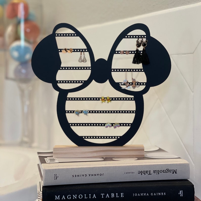 K&ouml;nnte beinhalten: Schwarzer Ohrringst&auml;nder aus Holz in Form einer Schleife mit Mickey-Maus-Ohren. Der St&auml;nder hat mehrere Reihen von L&ouml;chern f&uuml;r Ohrringe. Der St&auml;nder steht auf zwei B&uuml;chern, auf denen "Magnolia Table" von Joanna Gaines steht.