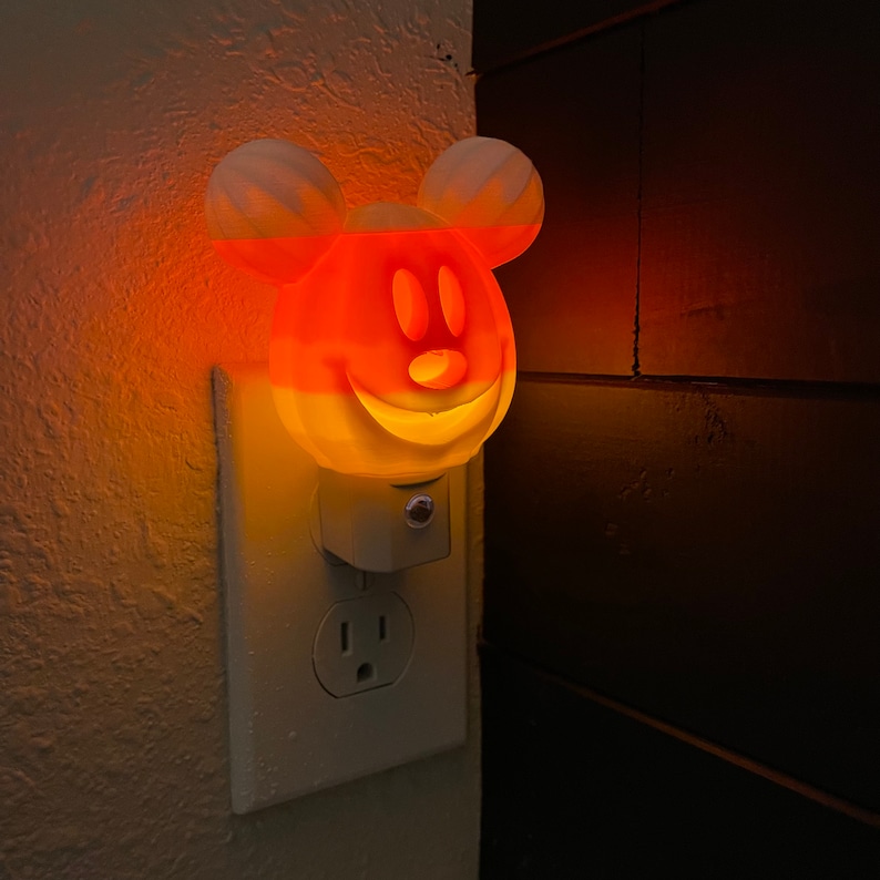 May include: A white and orange nightlight shaped like Mickey Mouse's head with a smiling face. The nightlight is plugged into an electrical outlet.